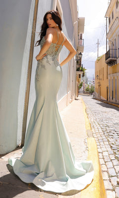 A woman in the Lace-Up Embroidered Bodice Long Prom Dress G1364, featuring a light blue, floor-length mermaid silhouette with beaded lace details, stands on a sunlit cobblestone street, facing away from the camera.