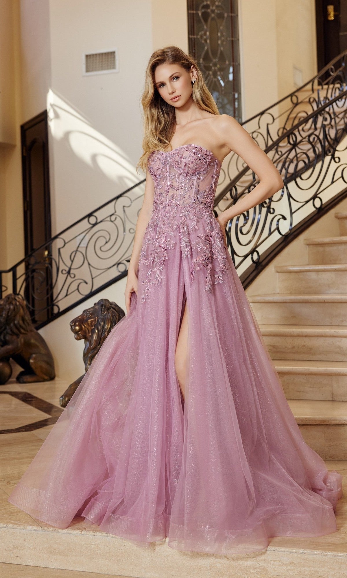 A woman stands on a staircase with ornate railing, wearing the Nox Anabel G1371 strapless long prom ball gown in lavender, featuring floral appliqués and a thigh-high slit.