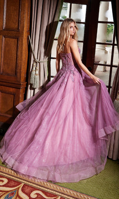 A woman in the Nox Anabel G1371 Strapless Long Prom Ball Gown with glitter and floral embroidery stands by a window, elegantly holding her dress and glancing over her shoulder.