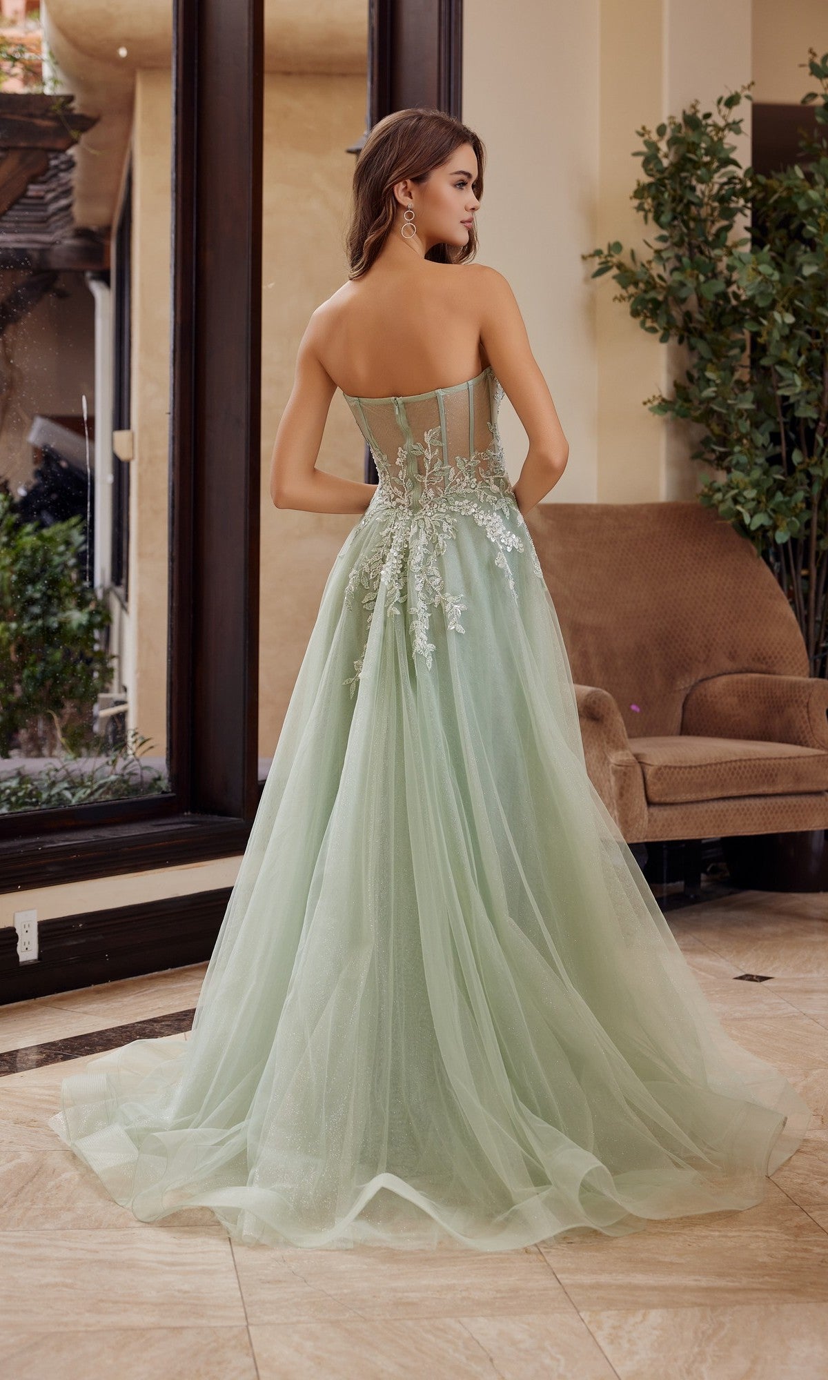 A woman in a Nox Anabel G1371 strapless, floor-length prom ball gown stands indoors, facing away from the camera.