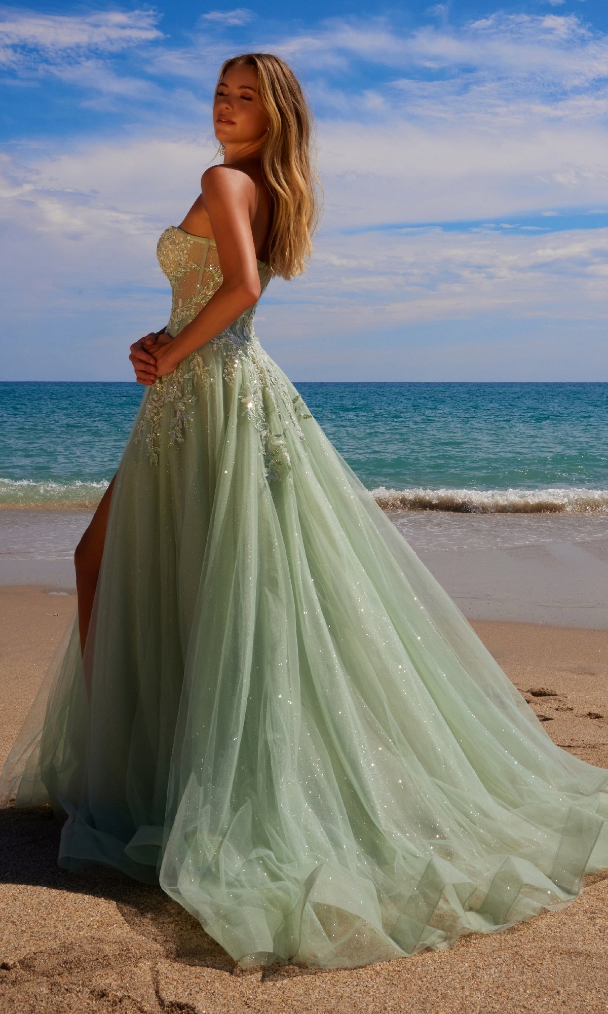 A woman stands on a sandy beach in the Nox Anabel G1371 Strapless Long Prom Ball Gown, her light green tulle dress shimmering against the ocean and blue sky.