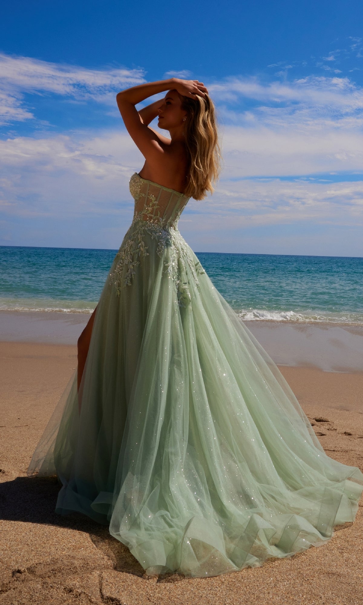 A woman in the Nox Anabel G1371 Strapless Long Prom Ball Gown stands on a sandy beach near the ocean, hand on her head, gazing at the horizon beneath a partly cloudy sky.