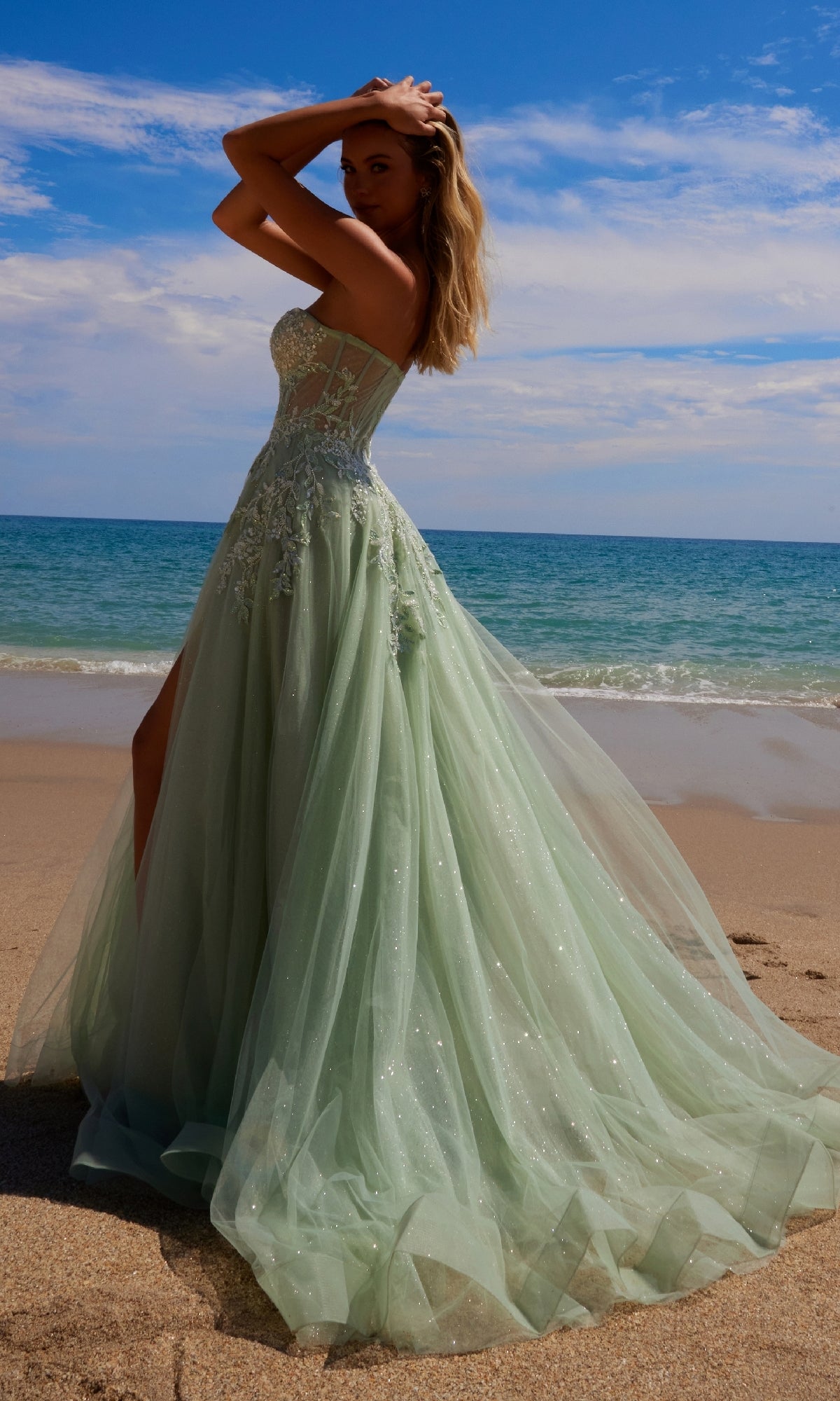 A woman stands on a sandy beach near the ocean, wearing the Nox Anabel G1371 strapless long prom ball gown with a thigh-high slit, her shimmering dress catching the light beneath a partly cloudy sky.