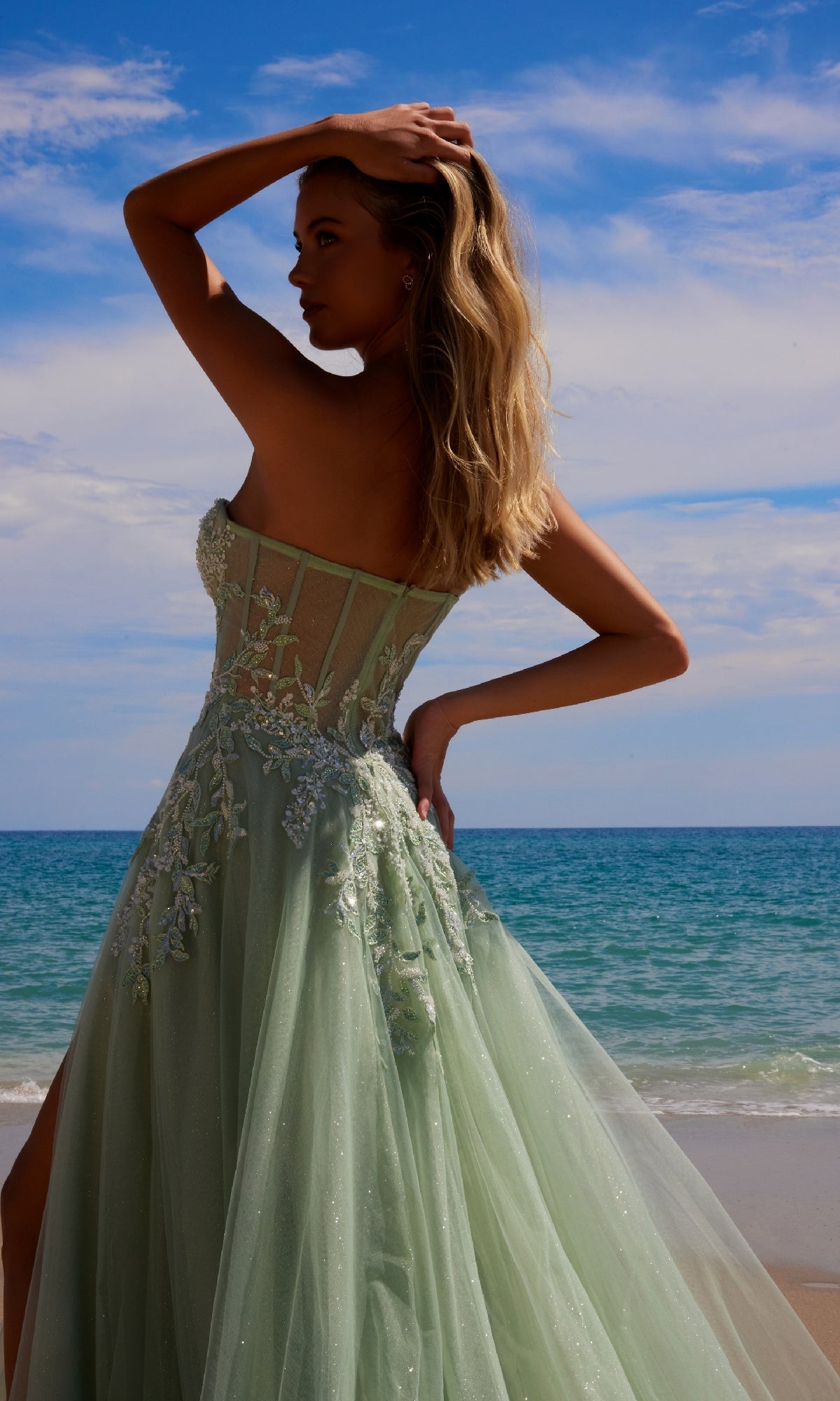 A woman wearing the Nox Anabel G1371 strapless long prom ball gown in pastel green embroidered lace stands on a beach with the ocean and blue sky in the background.