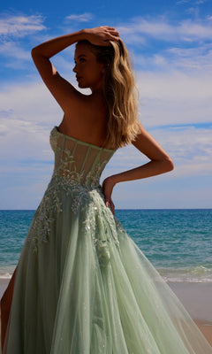 A woman wearing the Nox Anabel G1371 strapless long prom ball gown in pastel green embroidered lace stands on a beach with the ocean and blue sky in the background.