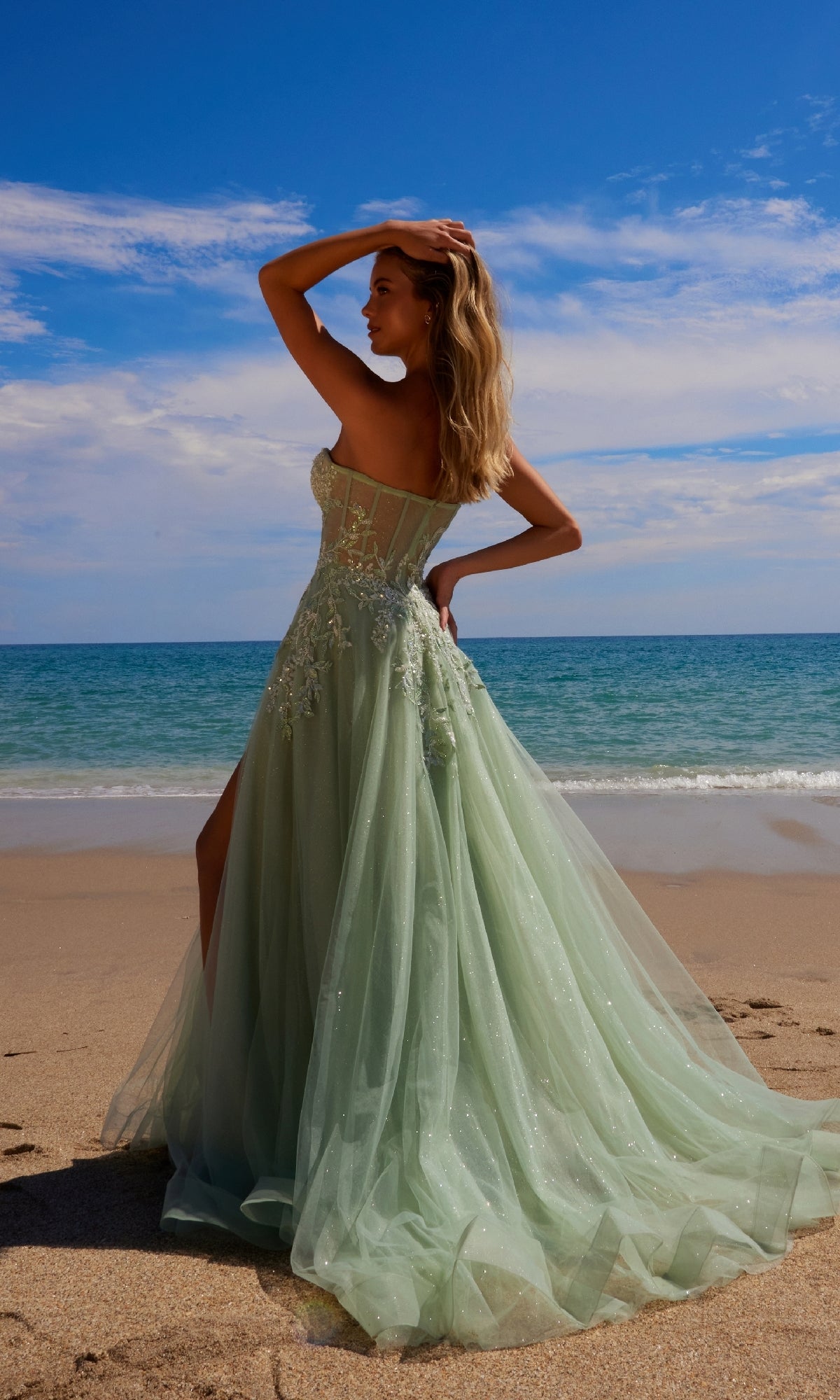 A woman in the Nox Anabel G1371 Strapless Long Prom Ball Gown stands on a sandy beach facing the ocean, one hand on her hip and the other touching her hair.