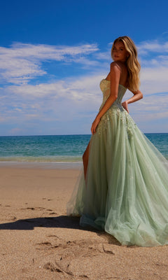 A woman in the Nox Anabel G1371 Strapless Long Prom Ball Gown stands on a sandy beach, facing the camera with the ocean and blue sky behind her.