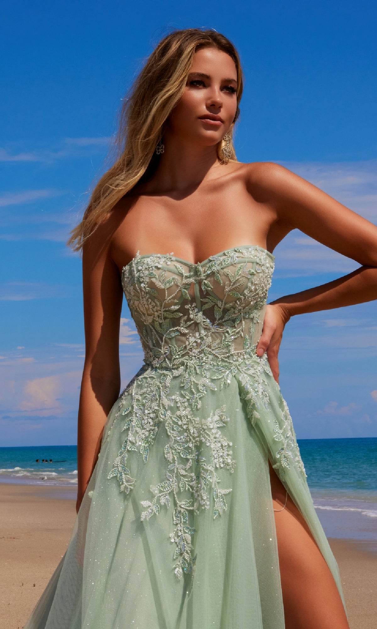 A woman stands on a sandy beach in the Strapless Long Prom Ball Gown: Nox Anabel G1371, her light green embellished dress shimmering against the ocean and blue sky backdrop.