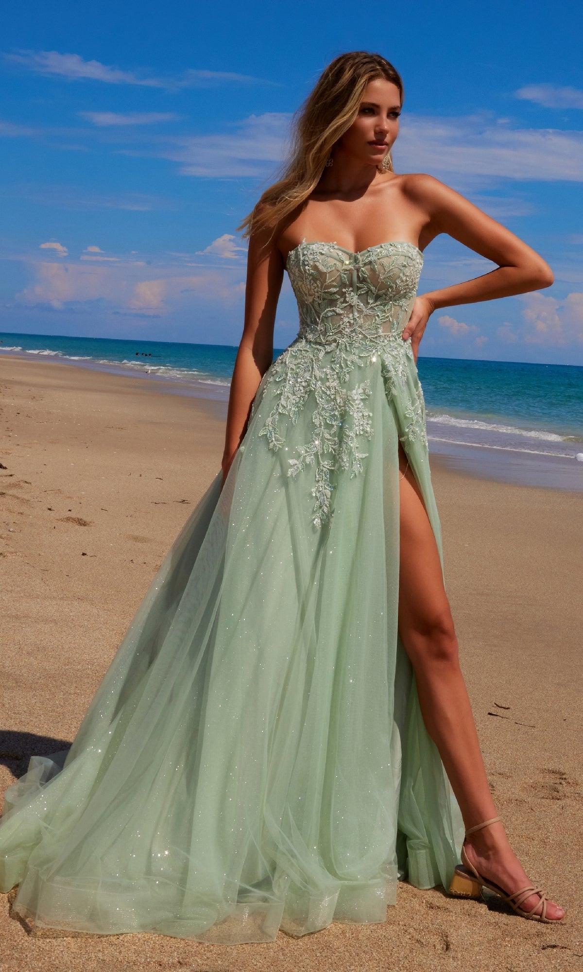 A woman in the Nox Anabel G1371 Strapless Long Prom Ball Gown with mint green floral embroidery and a high slit stands on a sandy beach by the ocean under a blue sky.
