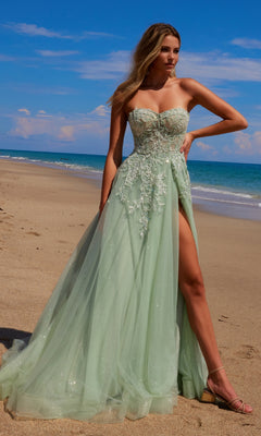 A woman in the Nox Anabel G1371 Strapless Long Prom Ball Gown with mint green floral embroidery and a high slit stands on a sandy beach by the ocean under a blue sky.
