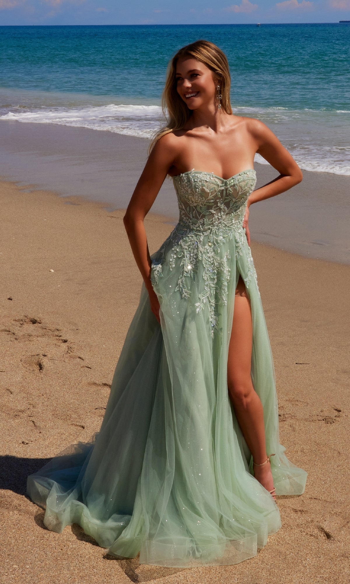 A woman in the Nox Anabel G1371 Strapless Long Prom Ball Gown with a thigh-high slit stands on a sandy beach near the ocean, smiling and posing.