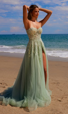 A woman wearing the Strapless Long Prom Ball Gown: Nox Anabel G1371, featuring light green embroidered lace, tulle layers, and a thigh-high slit, poses on a sandy beach with waves crashing behind her under a partly cloudy sky.