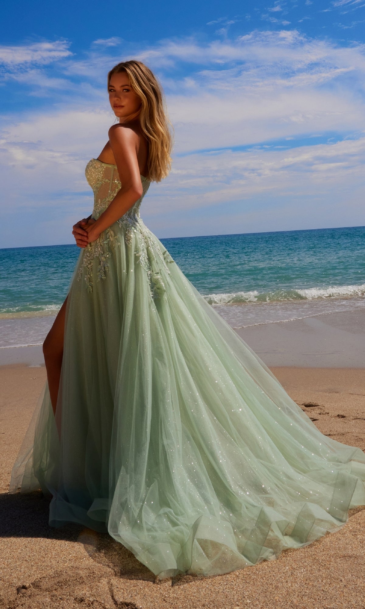 A woman wearing the Nox Anabel G1371 Strapless Long Prom Ball Gown with a flowing train stands on a sandy beach by the ocean under a partly cloudy sky.