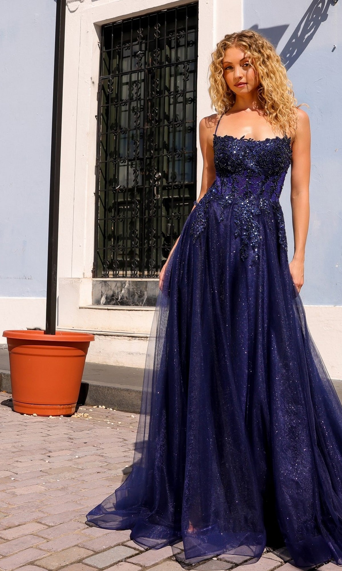 A woman wears the Embellished-Bodice Long A-Line Prom Dress G1405, standing on a paved area near a light blue building and a large empty plant pot.