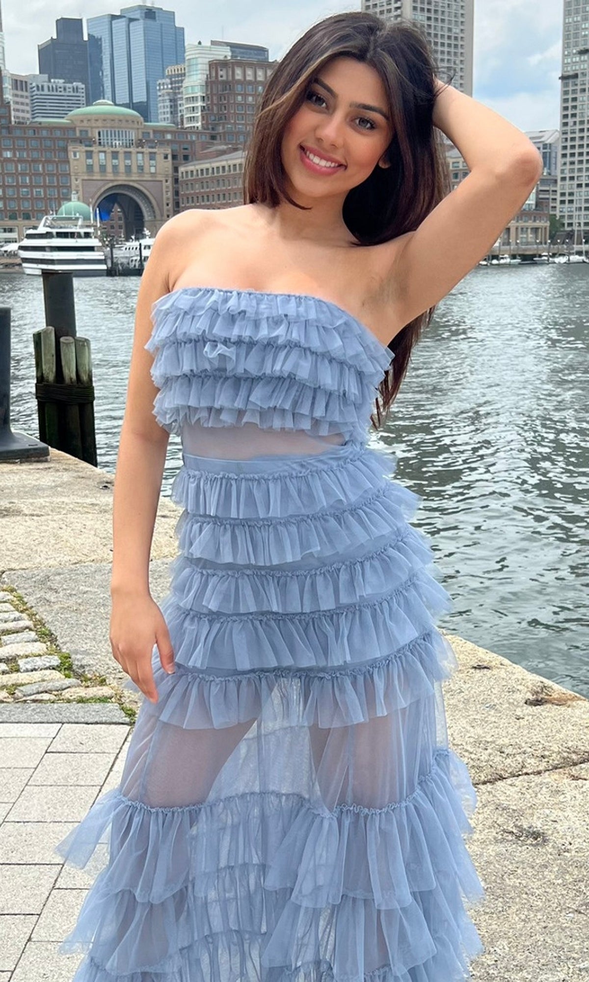 Wearing the Sheer Strapless Long Ruffled Prom Dress Georgia, a woman smiles at the camera by a waterfront with city buildings and a boat in the background.