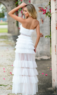 A woman stands outdoors before stone arches, wearing the Sheer Strapless Long Ruffled Prom Dress Georgia and glancing over her shoulder. Pink flowers bloom on a nearby vine.