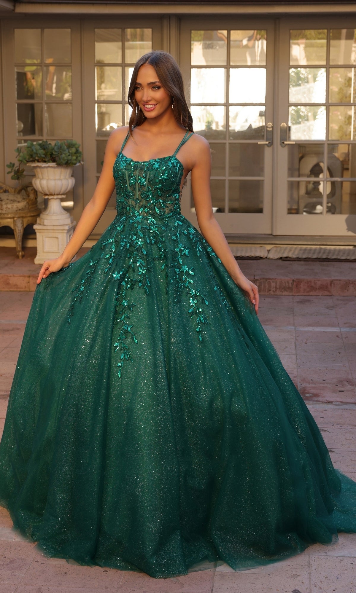 A woman stands outside in front of glass doors, wearing the Glitter-Tulle Long Prom Ball Gown H1271, a shimmering emerald green dress with floral embellishments and thin straps.