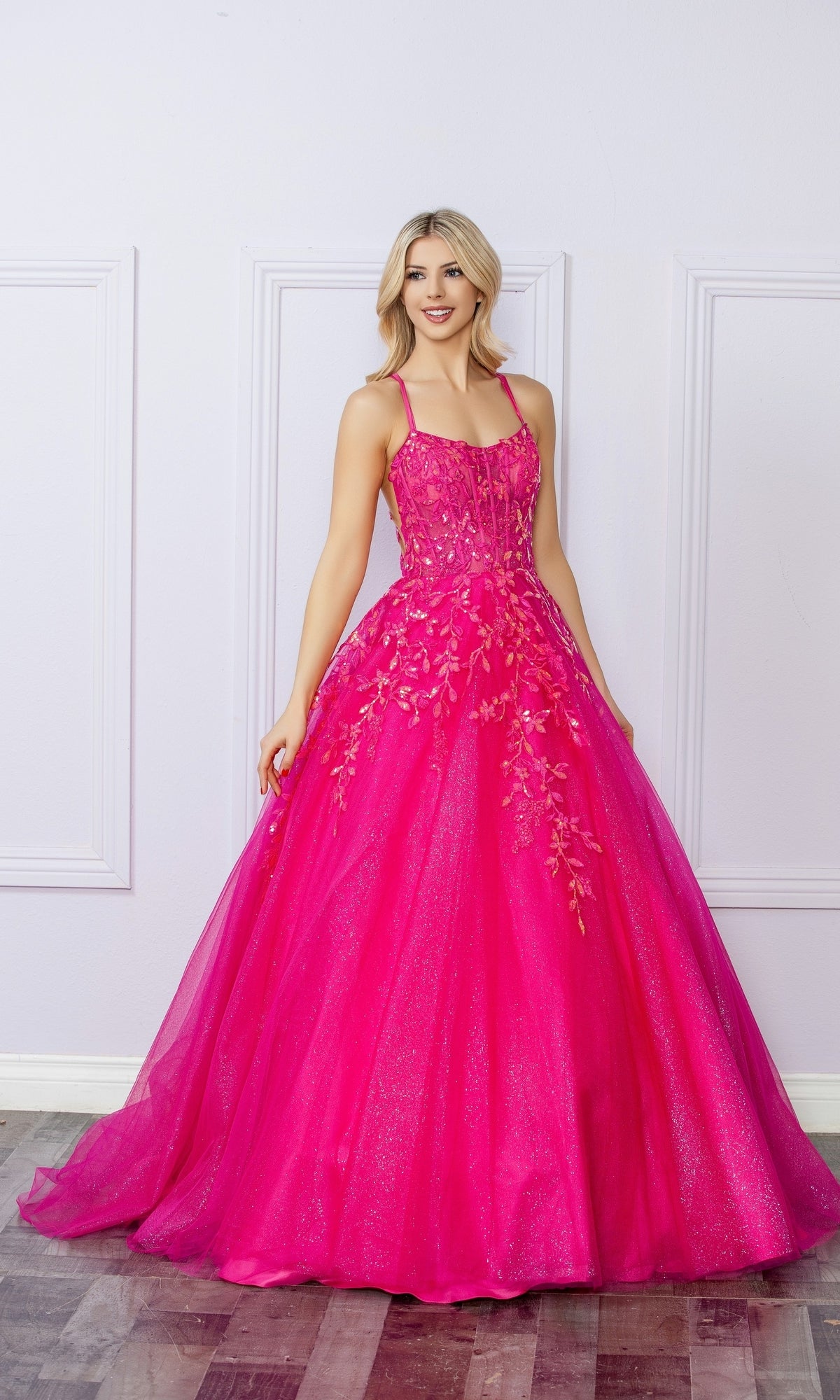 A woman poses indoors against a light-colored, paneled wall wearing the Glitter-Tulle Long Prom Ball Gown H1271, featuring bright pink glitter tulle, floral embellishments, and thin straps.