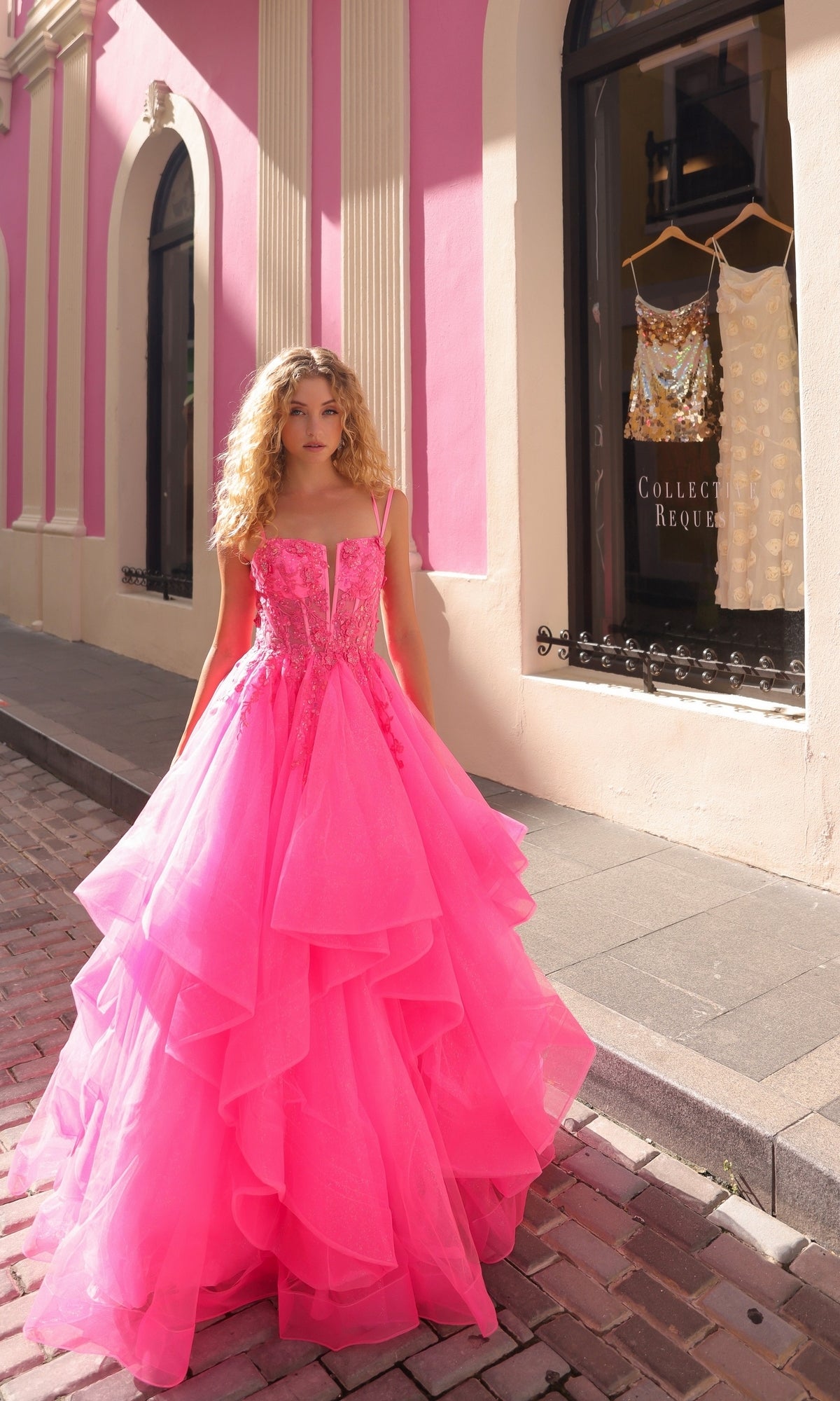 A woman with curly blonde hair wears the Lace-Up Long Tiered Prom Ball Gown H1351—a bright pink dress with tulle ruffles—while standing on a cobblestone street in front of a boutique displaying dresses in the window.