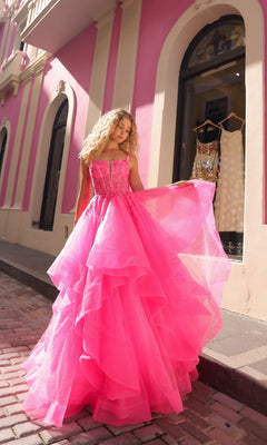 A woman wearing the Lace-Up Long Tiered Prom Ball Gown H1351, a voluminous bright pink tulle dress with dramatic ruffles, stands on a cobblestone sidewalk in front of a pink and white building with arched windows.