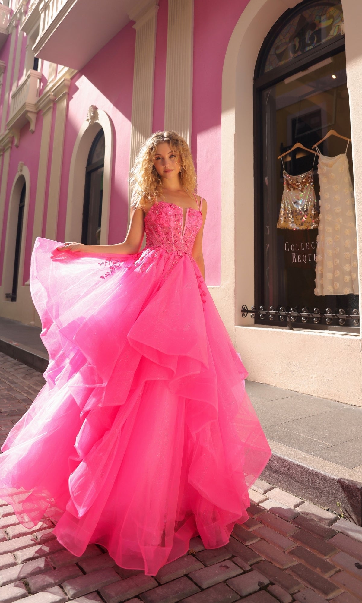 A woman poses in front of a pink storefront, wearing the Lace-Up Long Tiered Prom Ball Gown H1351 with tulle ruffles and a lace-up corset back.