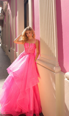 A woman wearing the Lace-Up Long Tiered Prom Ball Gown H1351 with bright pink tulle ruffles stands against a sunlit building with pink walls and tall windows.
