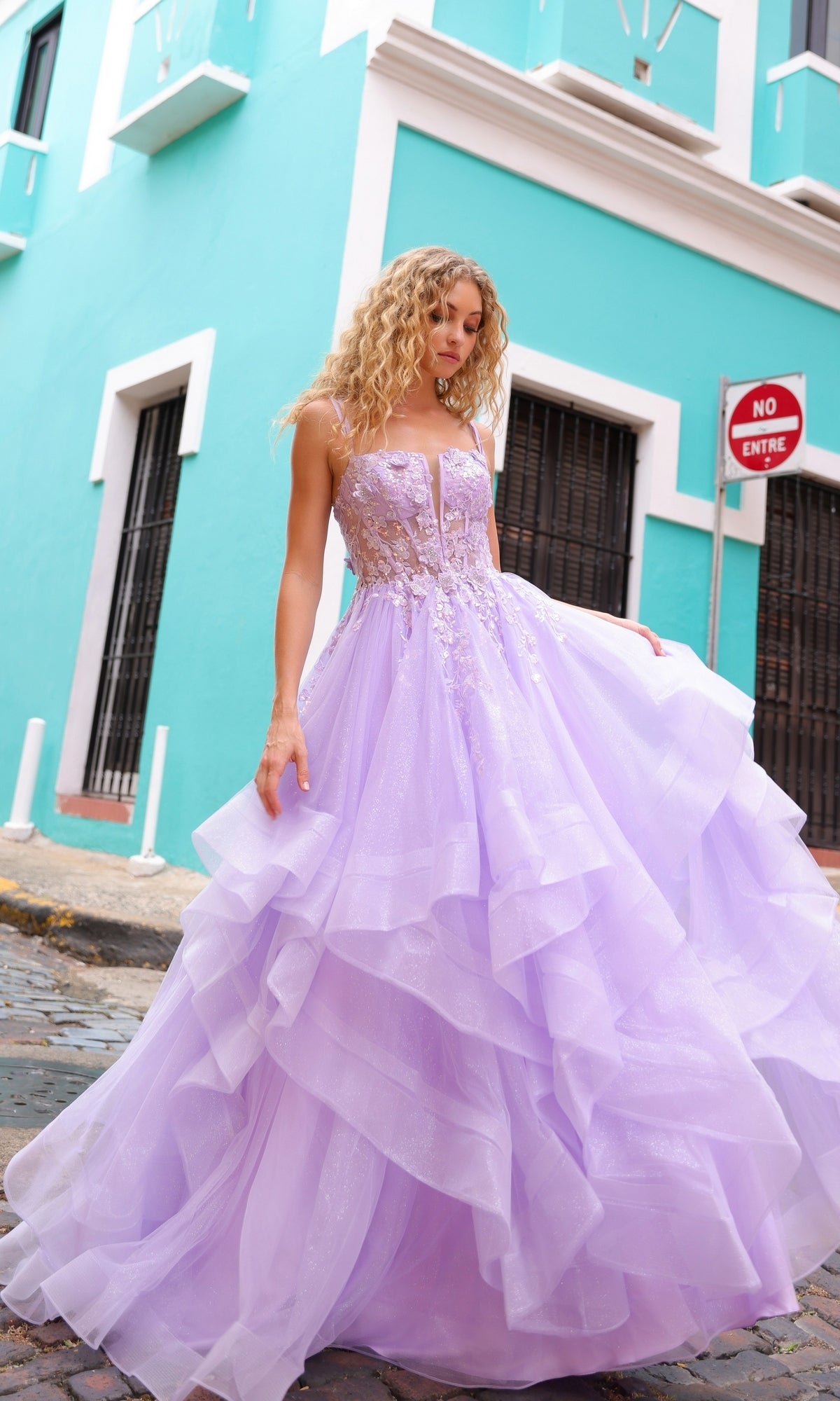 A woman wearing the Lace-Up Long Tiered Prom Ball Gown H1351 with tulle ruffles stands on a cobblestone street before a turquoise building displaying a 