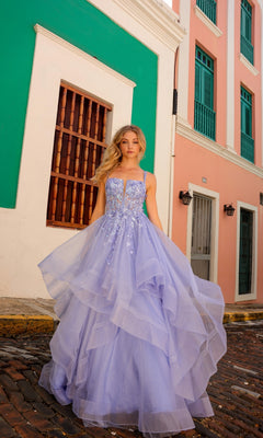 A woman wearing the Lace-Up Long Tiered Prom Ball Gown H1351, a flowing lavender dress with tulle ruffles, stands on a cobblestone street amid pastel buildings with green shutters and a wooden window.