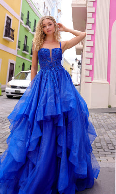 A woman wearing the Lace-Up Long Tiered Prom Ball Gown H1351 in bright blue, featuring tulle ruffles and a corset back, stands on a cobblestone street with colorful buildings and a white car behind her.