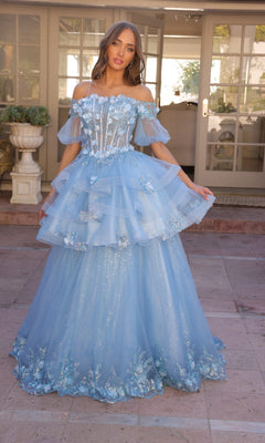 A woman stands outdoors in front of glass doors wearing the Nox Anabel Removable-Skirt Formal Ball Gown H1360, a light blue off-the-shoulder dress featuring floral details, a layered tulle skirt, and sheer overlay.