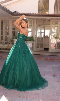 A woman stands outdoors before glass doors, wearing the Embellished Hunter Green Long Prom Ball Gown H1464 with sparkling sequins and a flowing A-line silhouette, arms raised above her head.