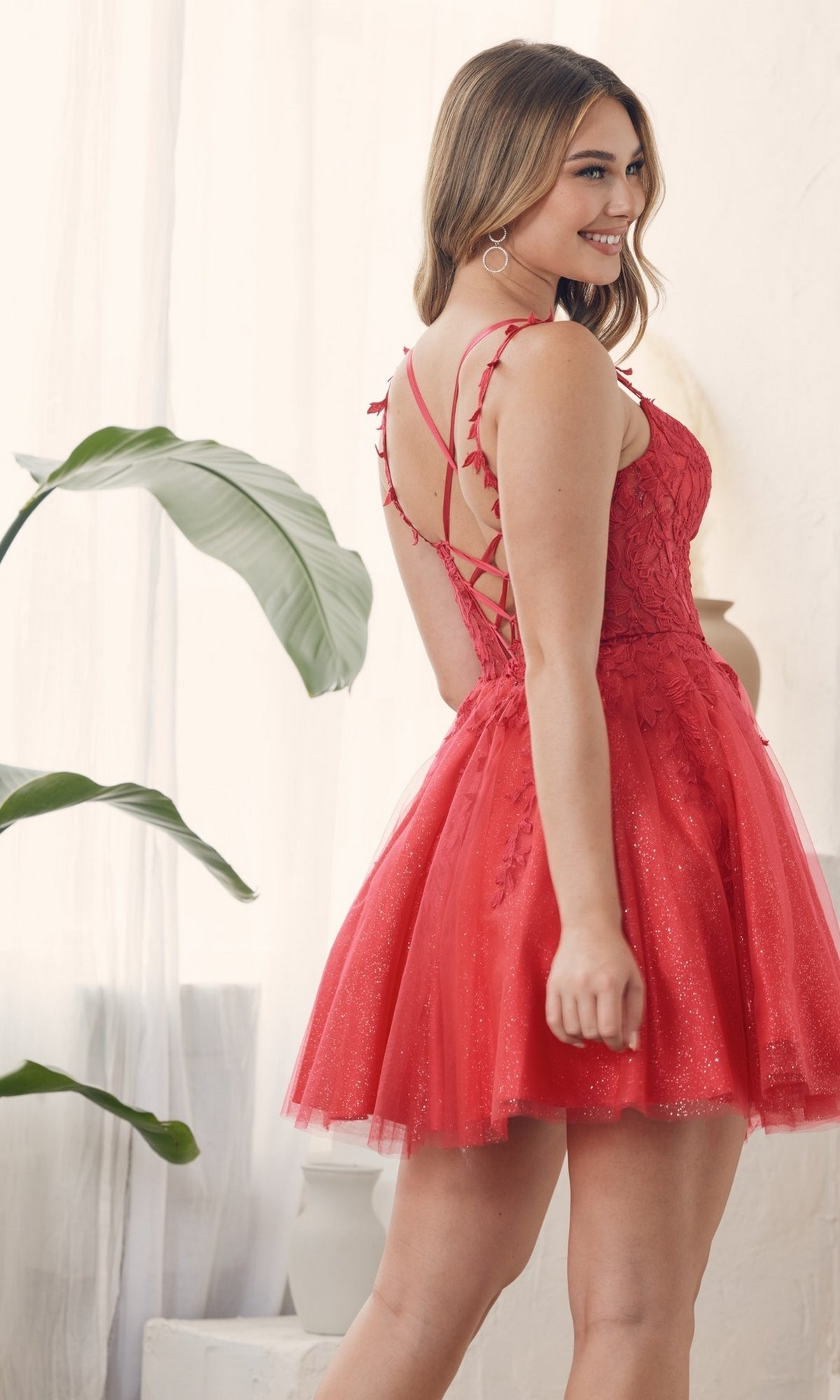 A woman in the Sheer-Waist Short Babydoll Homecoming Dress H784, featuring a lace-up back and sleeveless red embroidered design, stands indoors smiling over her shoulder. A green plant decorates the background.