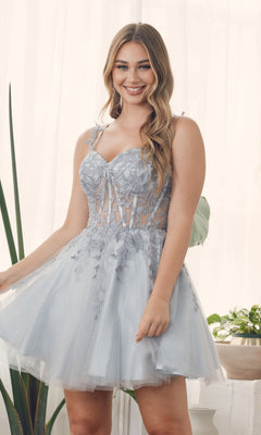 A woman wears the Sheer-Waist Short Babydoll Homecoming Dress H784—an embroidered light blue lace and tulle dress—standing indoors, smiling at the camera with plants in the background.