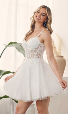 A woman stands indoors, smiling, in the Sheer-Waist Short Babydoll Homecoming Dress H784—an embroidered white lace and tulle dress with floral details.
