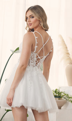 A woman stands indoors near plants in the Sheer-Waist Short Babydoll Homecoming Dress H784, a short, white, sleeveless dress with leaf embellishments and a lace-up back for an elegant and affordable homecoming look.