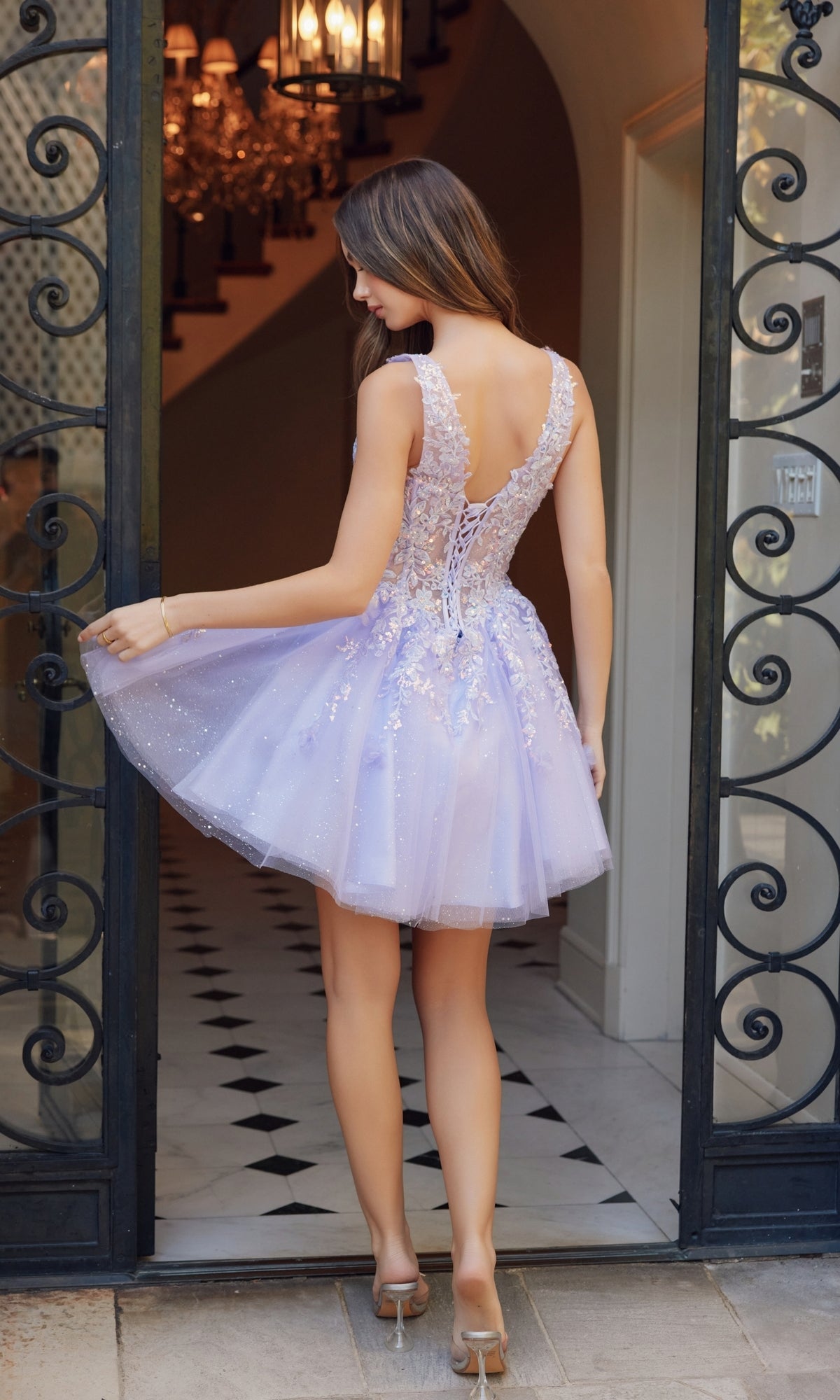 A woman in the Embroidered-Bodice Short A-Line Hoco Dress H846 stands by an open wrought iron gate, holding out one side of her light purple tulle skirt with its embroidered-lace sweetheart bodice.