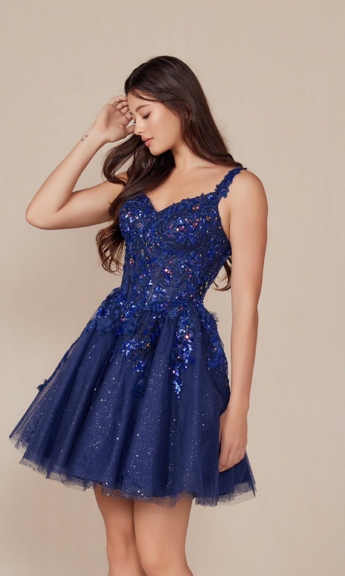 A woman wearing the Embroidered-Bodice Short A-Line Hoco Dress H846, featuring a dark blue sleeveless sequin top and tulle skirt, stands against a beige background, looking down and touching her hair.