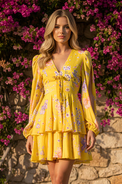 A woman wears the Buttercup Yellow Floral Print Casual Dress: Hannah, featuring ruffled A-line tiers, while standing by a stone wall and blooming pink flowers—an ideal choice for a wedding guest.