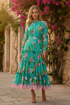 A woman stands outdoors on a stone path, wearing the Tropical Tea Length Casual Dress: Hazel in tropical teal. The dress features long sleeves, a belted waist, and is surrounded by pink flowers and greenery.