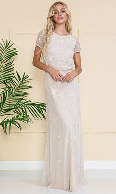A woman in the Blouson Long Formal Dress: Amelia Couture IN004, featuring delicate floral embellishments, stands indoors beside a green leafy plant and a cream-colored wall.