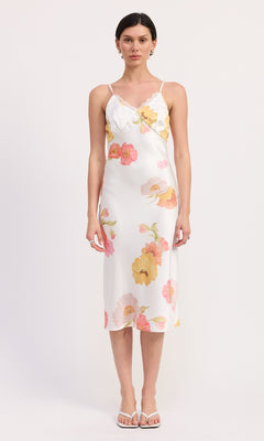 A woman faces forward in a V-Neck Floral Print Midi Slip Dress with spaghetti straps and white sandals, set against a plain background.