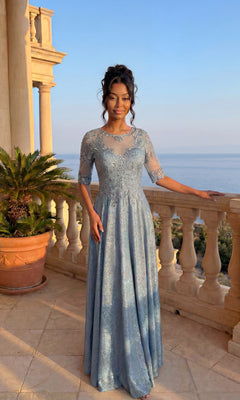 A woman in the blue Long Formal Dress: Chicas J006 stands on a stone balcony with a potted plant, overlooking the ocean under a clear sky.