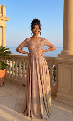 A woman stands on a terrace by the coast, wearing the Chicas J006 Long Formal Dress in rose gold, featuring lace embroidery and sheer sleeves.