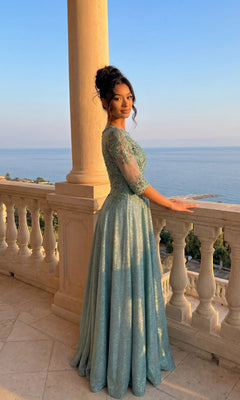 A woman in the sage Long Formal Dress: Chicas J006 with lace embroidery stands on a balcony at sunset, surrounded by stone columns and balustrades, overlooking the sea.