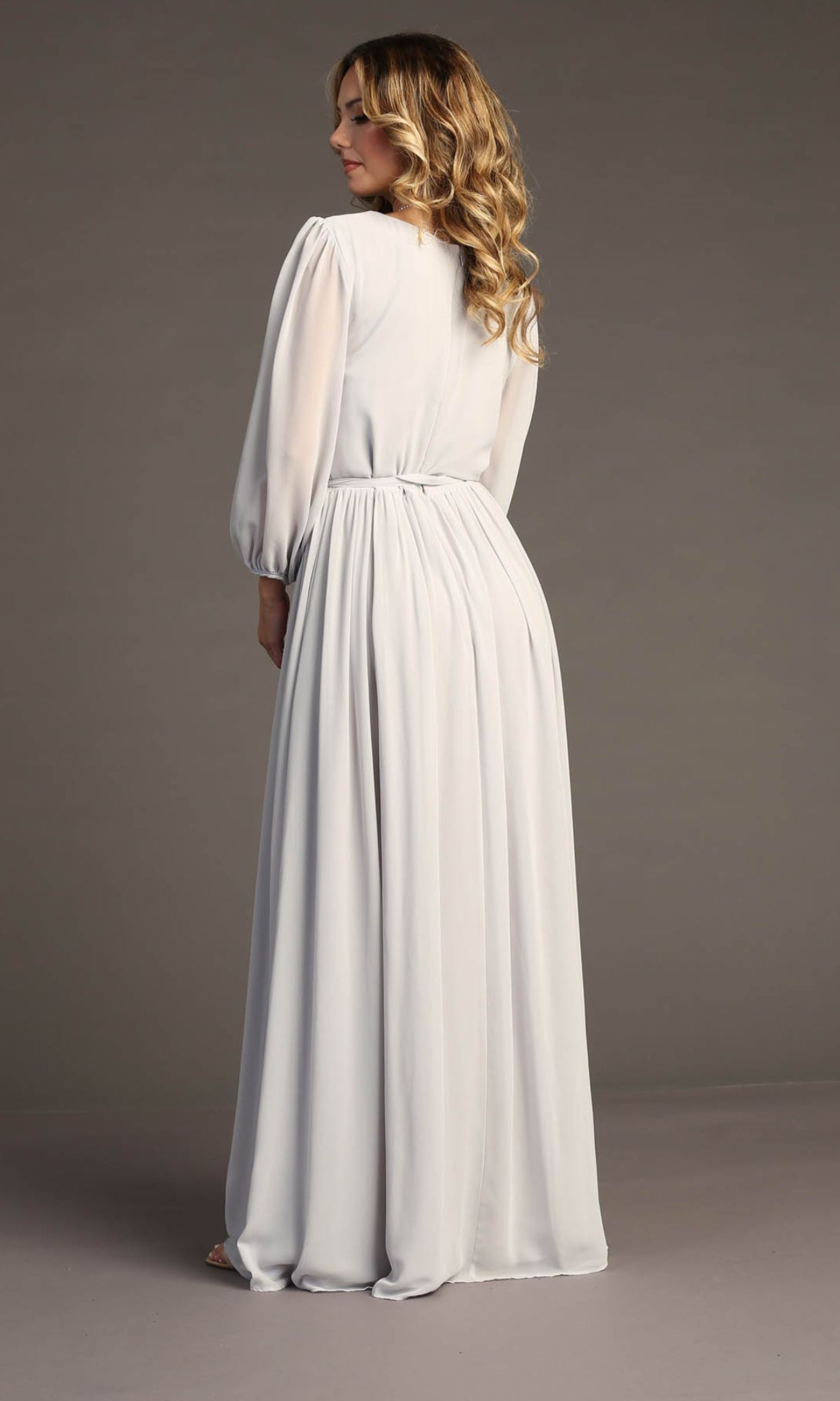 A woman with wavy hair stands with her back to the camera, modeling the Chicas J012B Long-Sleeve Long Formal Dress in light gray, featuring gathered fabric, against a plain background.