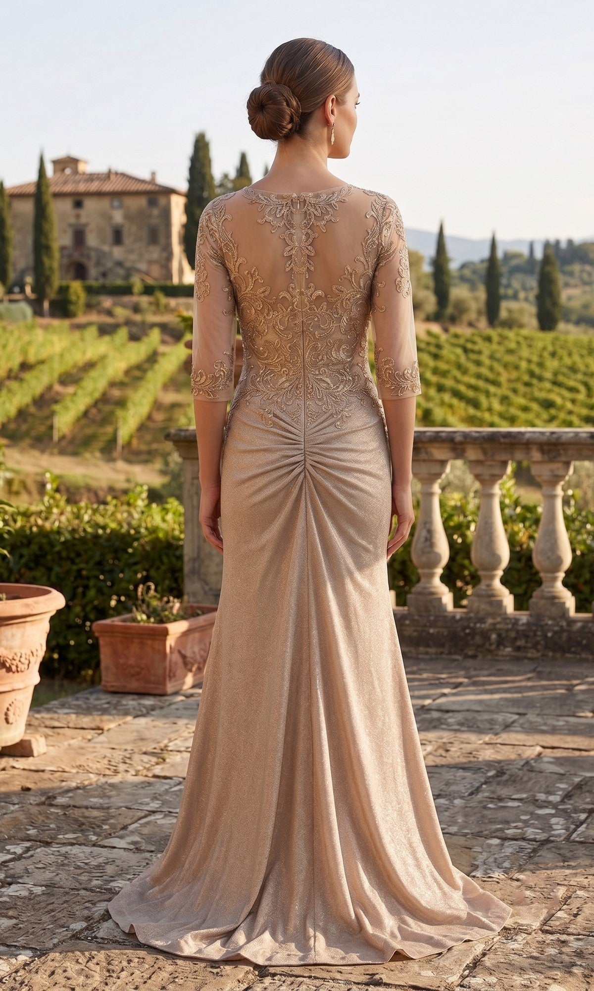 A woman wearing the Long Prom Dress: Chicas J046—an elegant, floor-length formal gown featuring a sheer bodice and delicate gold embroidery—stands outdoors on a stone patio, facing away as she overlooks a vineyard and historic building.
