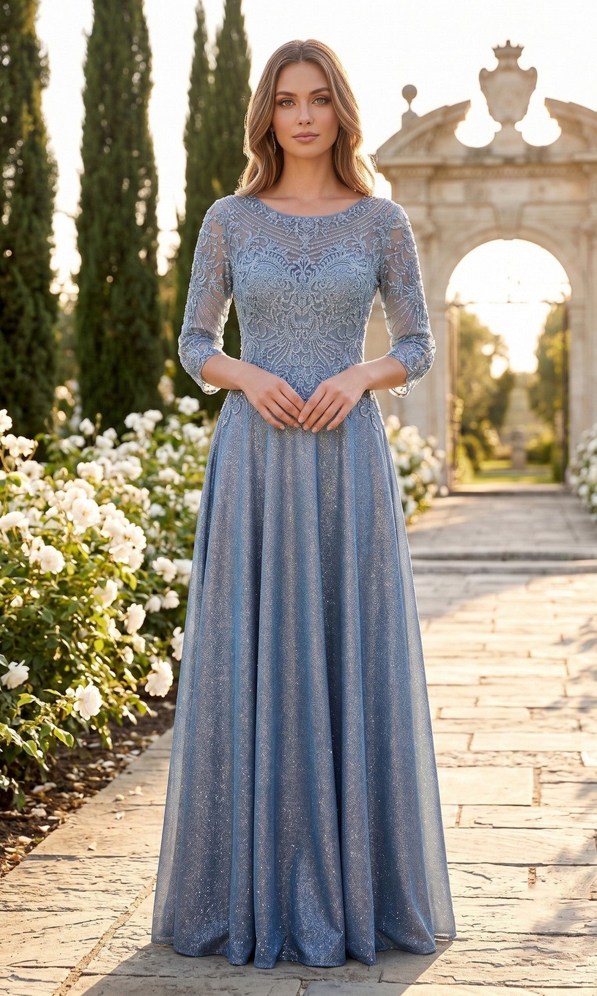 A woman stands on a garden path wearing the Chicas J052 long formal dress in blue—a modest gown with three-quarter sleeves and delicate lace. White flowers and greenery line the walkway, with stone arches in the background.