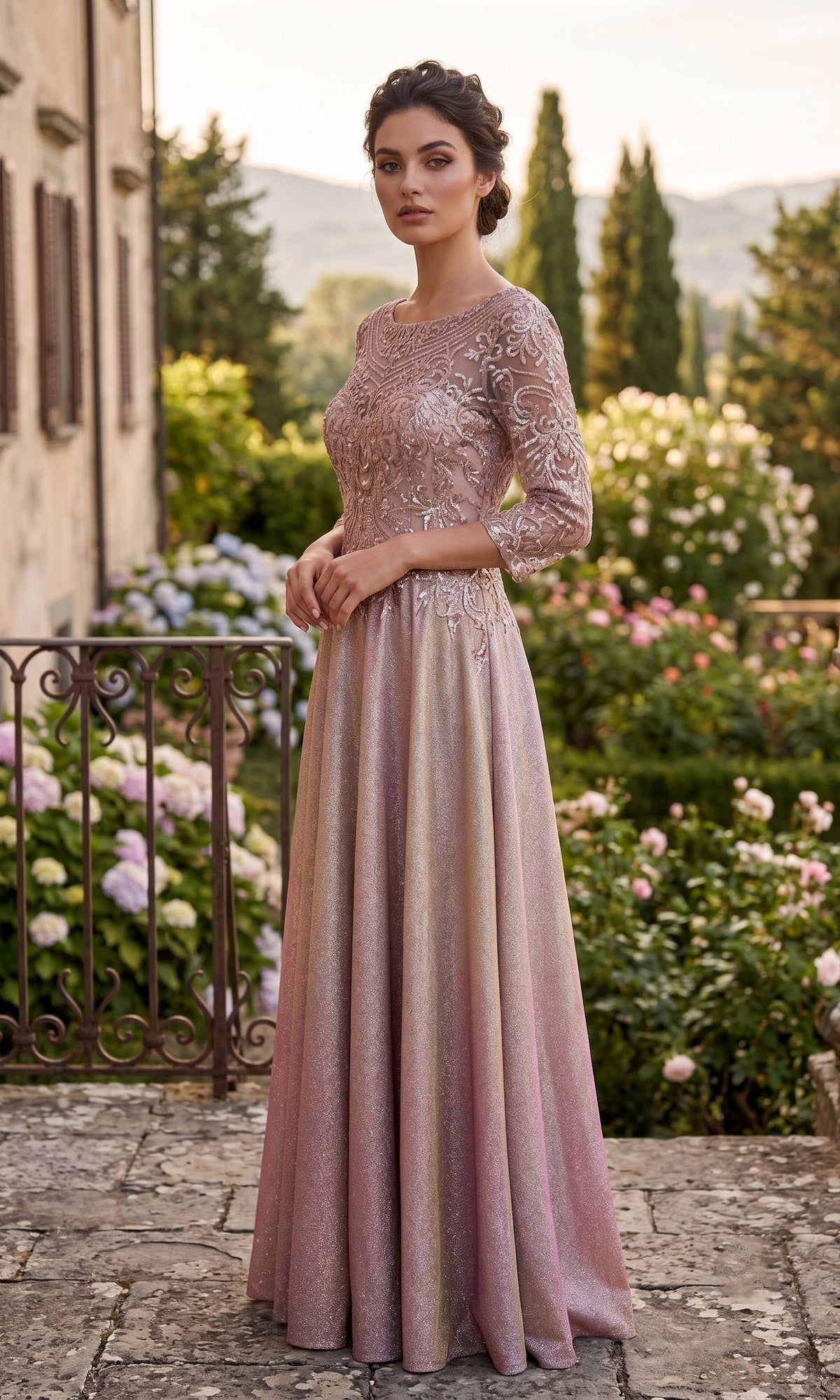 A woman wearing the Long Formal Dress: Chicas J052—a long, light pink lace gown featuring an elegantly embroidered bodice—stands outdoors on a stone terrace, surrounded by flowering bushes and lush greenery.