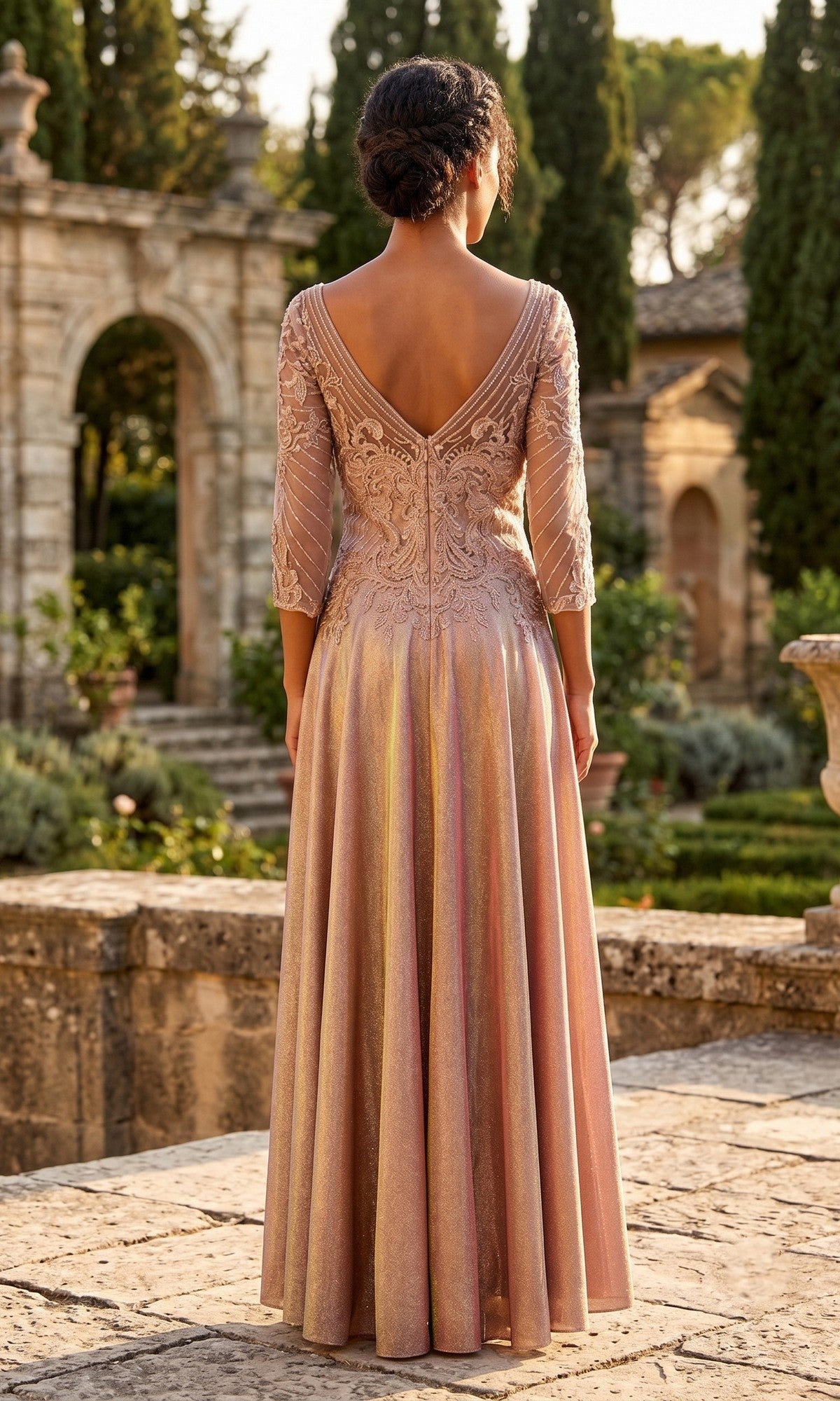 A woman in the Long Formal Dress: Chicas J052—a light pink, floor-length gown featuring an embroidered bodice and lace sleeves—stands outdoors on stone paving, facing away, with greenery and stone arches in the background.