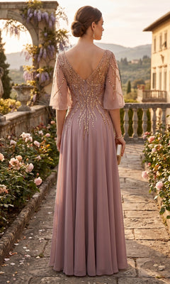 A woman in the Long Prom Dress: Chicas J064—a mauve mother-of-the-bride gown featuring beaded details—stands on a stone path in a garden, holding a clutch, with flowers and historic buildings in the background.